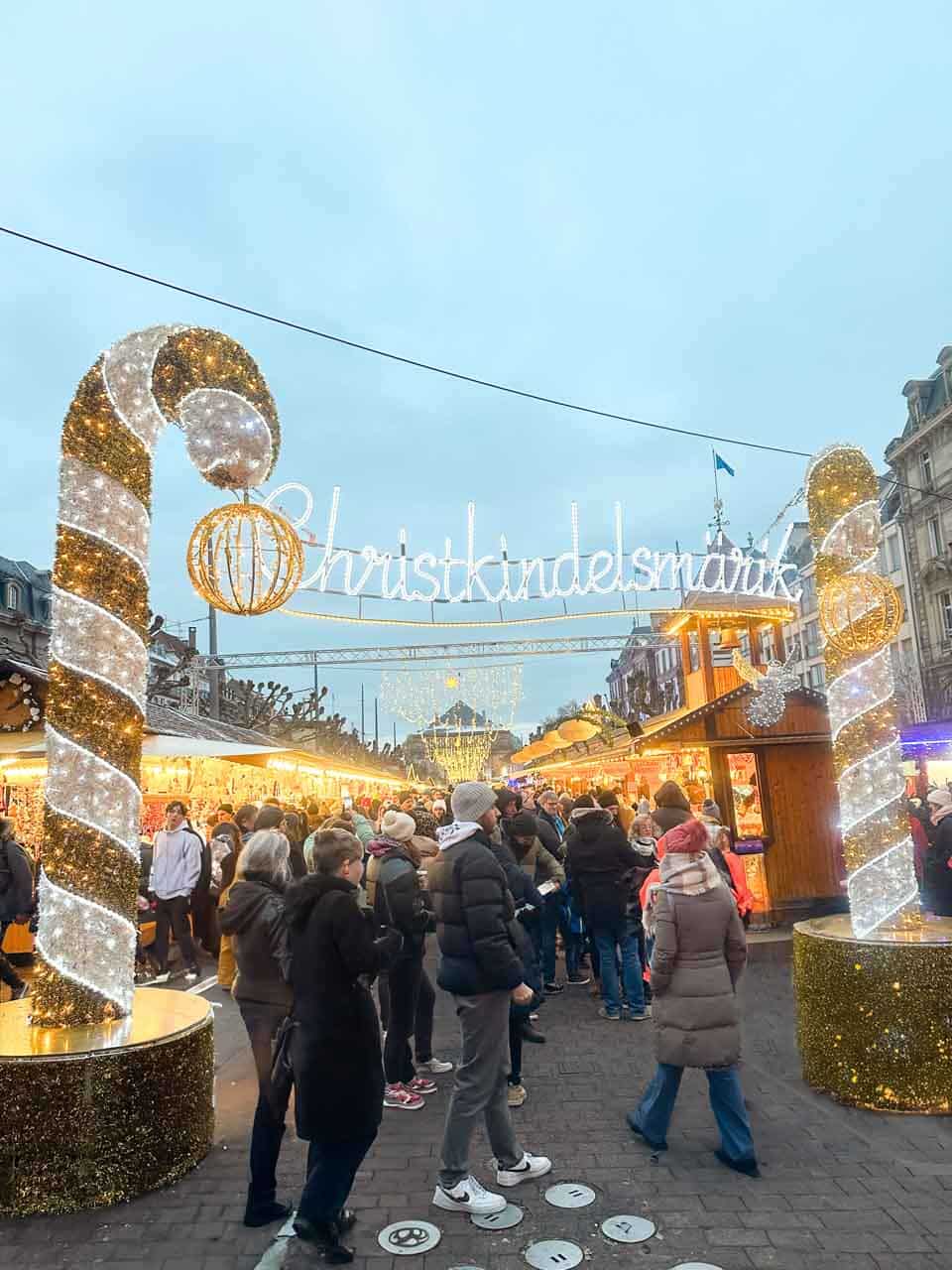 Visitors at the entrance of the Christkindelsm&auml;rik Christmas Market in Strasbourg, framed by shimmering candy cane arches with the market's name glowing above in festive lights