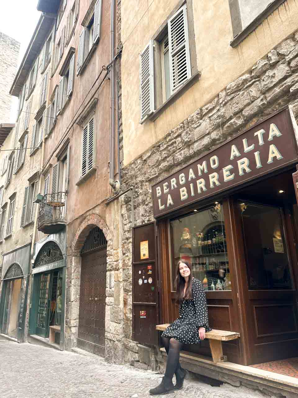 A smiling woman sitting on a wooden bench outside a bar named Bergamo Alta La Birreria