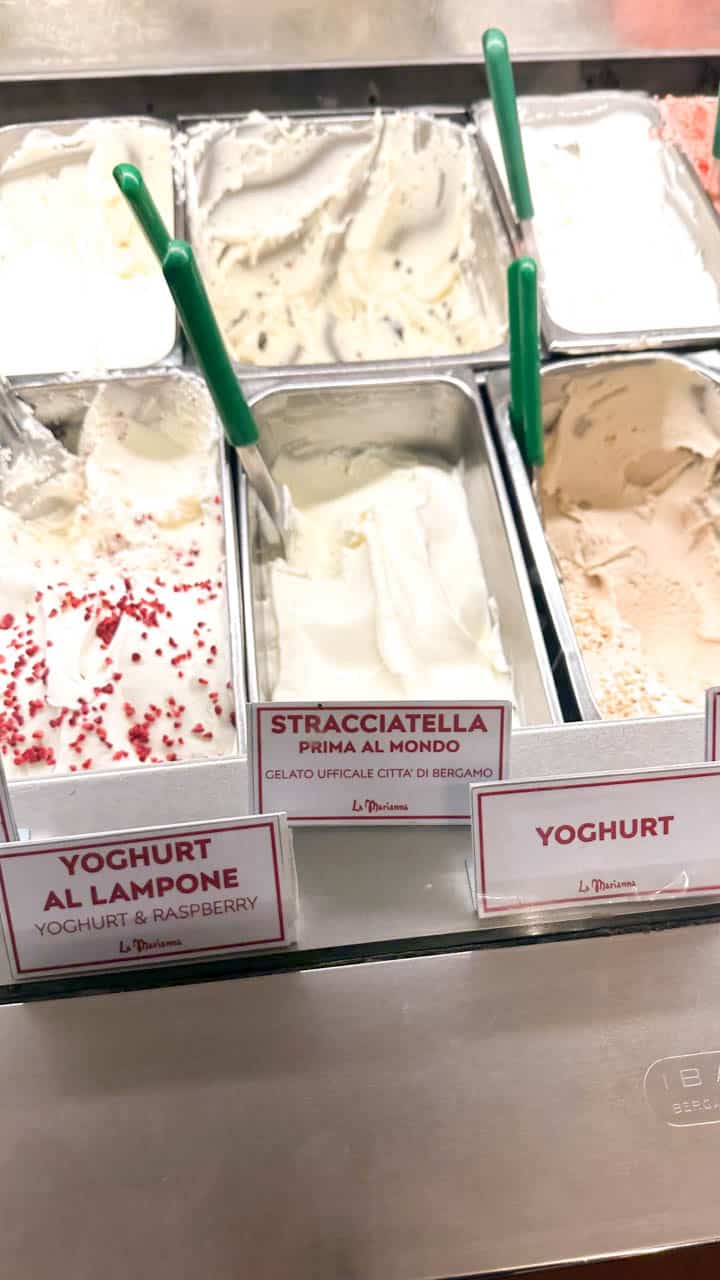 Several trays of gelato displayed in a freezer, with flavour labels indicating 'Yoghurt', 'Stracciatella', and 'Yoghurt al Lampone' at the La Marianna gelateria in Bergamo, Italy