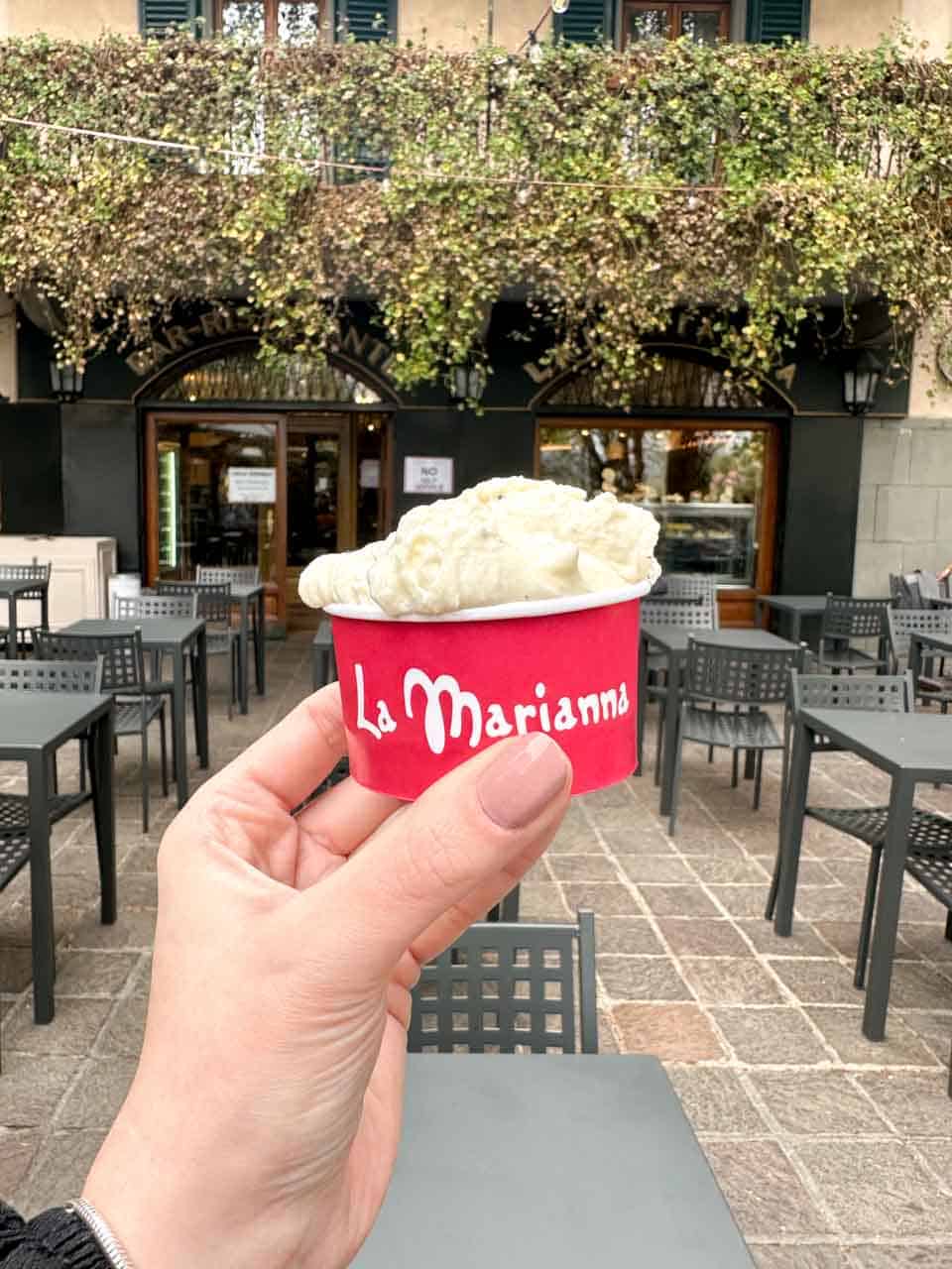 A close-up of a hand holding a small red cup of stracciatella gelato, with an outdoor seating area of a café and a vine-covered building in the background