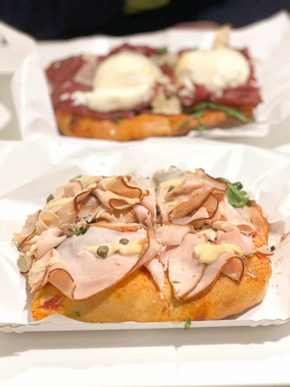 Two slices of pizza topped with cured meats and cheese, served on white paper trays at Il Fornaio in Bergamo, Italy