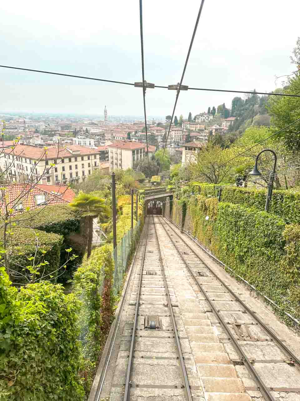 The view from a funicular railway showing tracks descending a hill with a panoramic view of Bergamo cityscape below