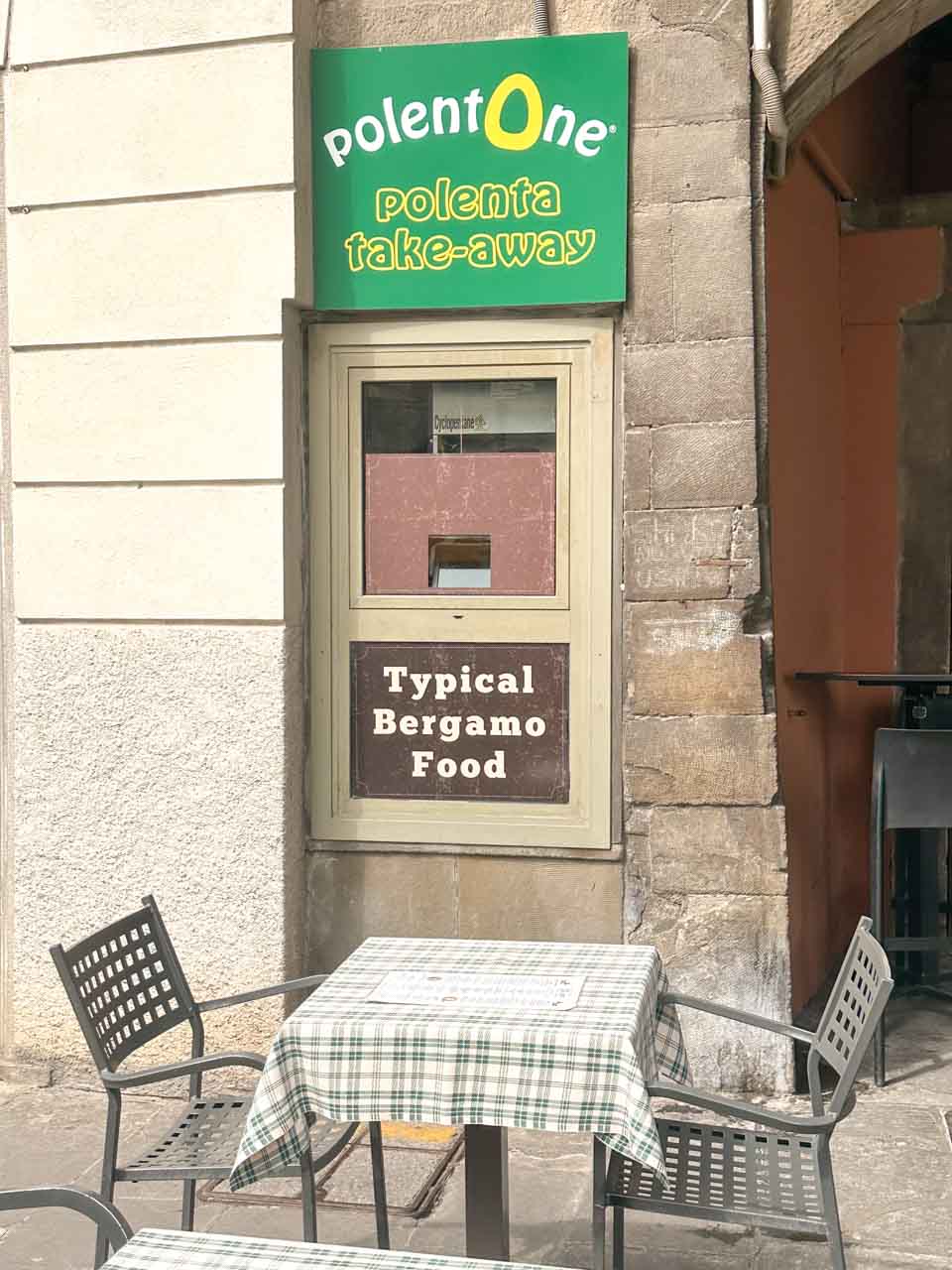 A street view of 'polentOne take-away' with a bright green sign, a table with a checkered tablecloth in front, and a sign reading 'Typical Bergamo Food'