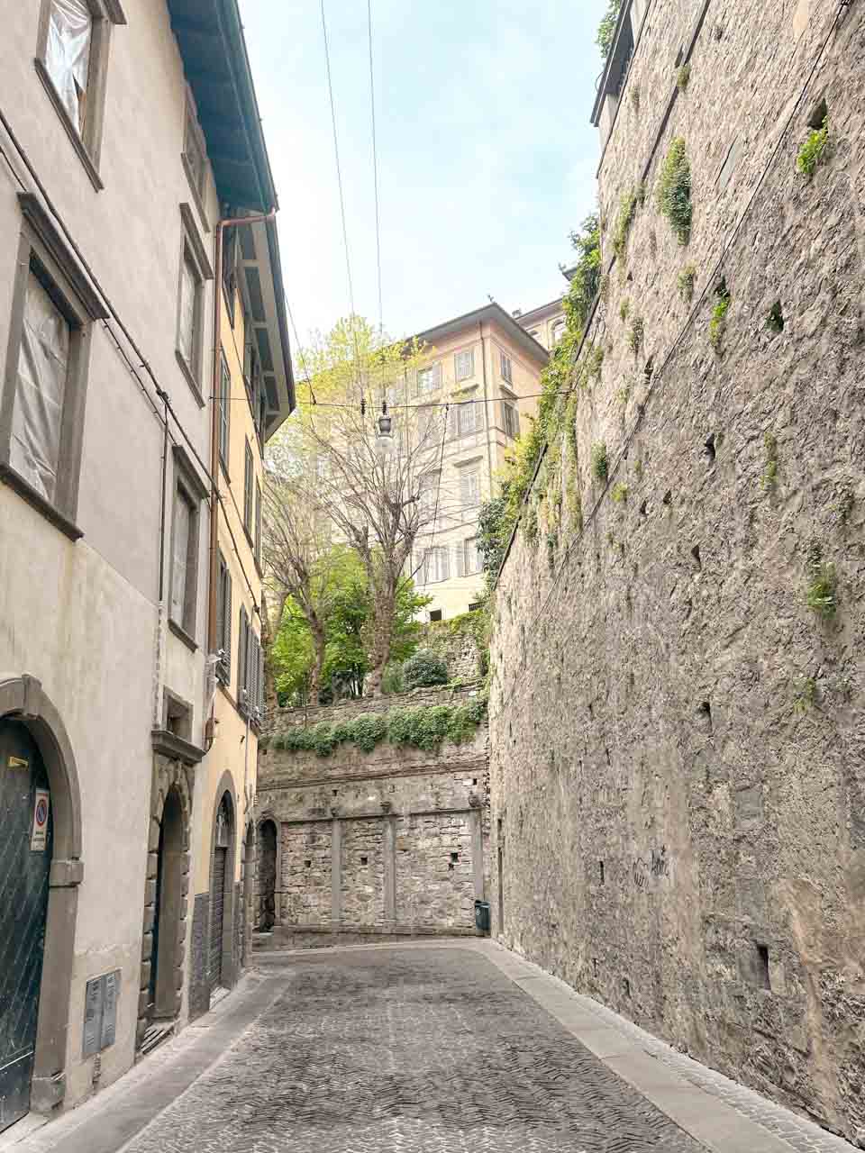 A narrow cobbled street in Bergamo, flanked by old buildings with a high stone wall on one side, with lush greenery visible above the wall