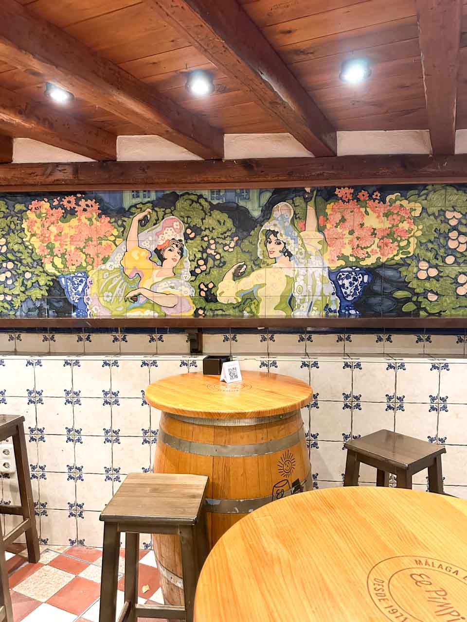 The interior of Bodega El Pimpi in Malaga, Spain, with a decorative tile mural on the wall and wooden barrels as tables