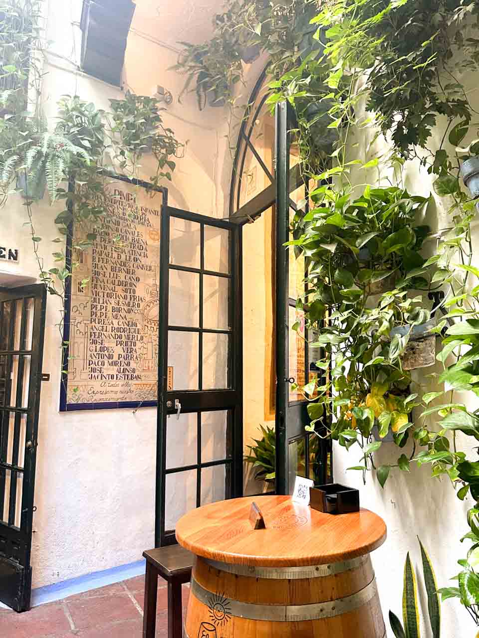 A corner of Bodega El Pimpi with green plants climbing the walls, a wooden barrel table, and a historic poster framed on the wall