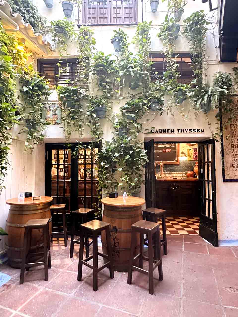 A charming courtyard of Bodega El Pimpi with wooden barrel tables and stools, surrounded by greenery and the facade of an old building