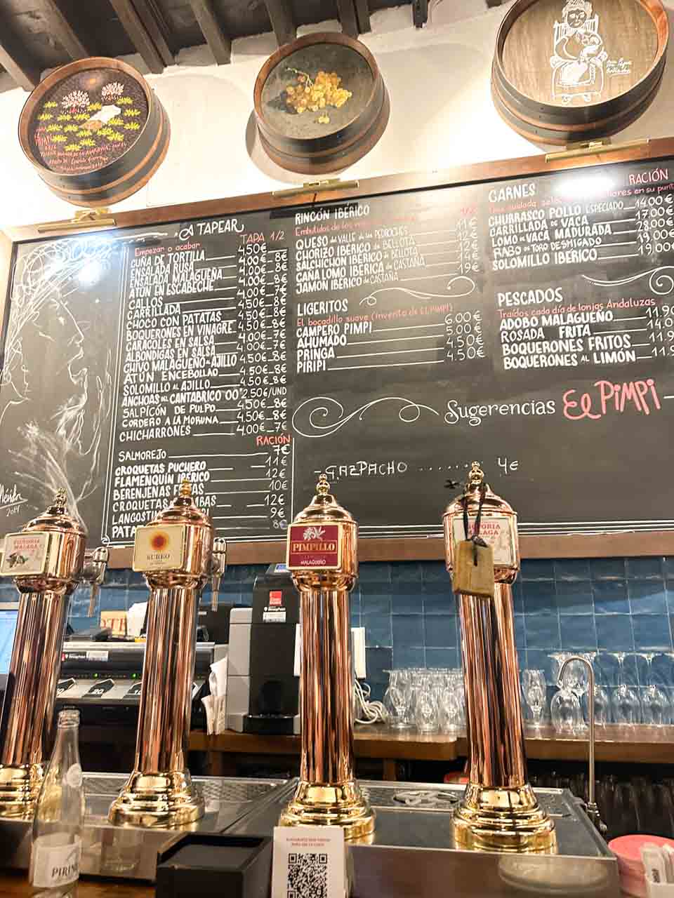 A chalkboard menu showcasing a variety of Spanish dishes and prices, above shiny beer taps and a bar counter at Bodega El Pimpi