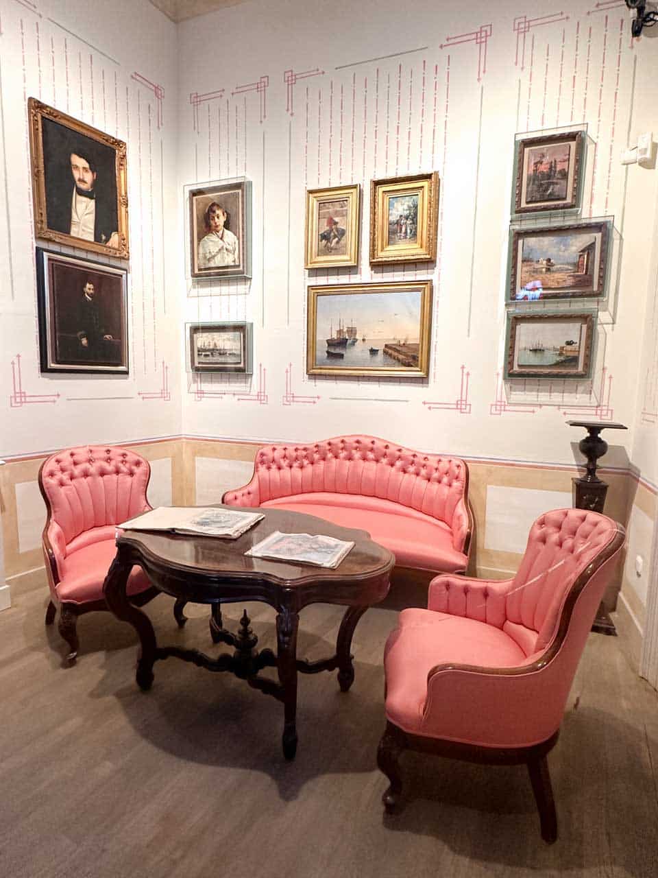 A room inside the Picasso Birthplace Museum with pink tufted sofas and a wooden table, surrounded by framed paintings hung on a white wall