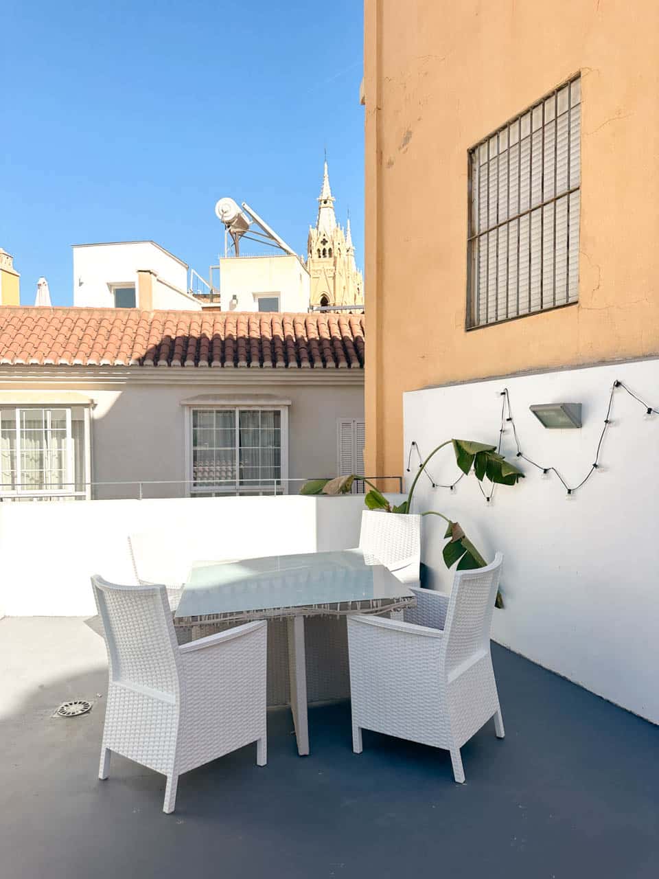 A rooftop terrace with white wicker chairs and a glass table, offering a view of Malaga Cathedral&rsquo;s spire against a clear sky