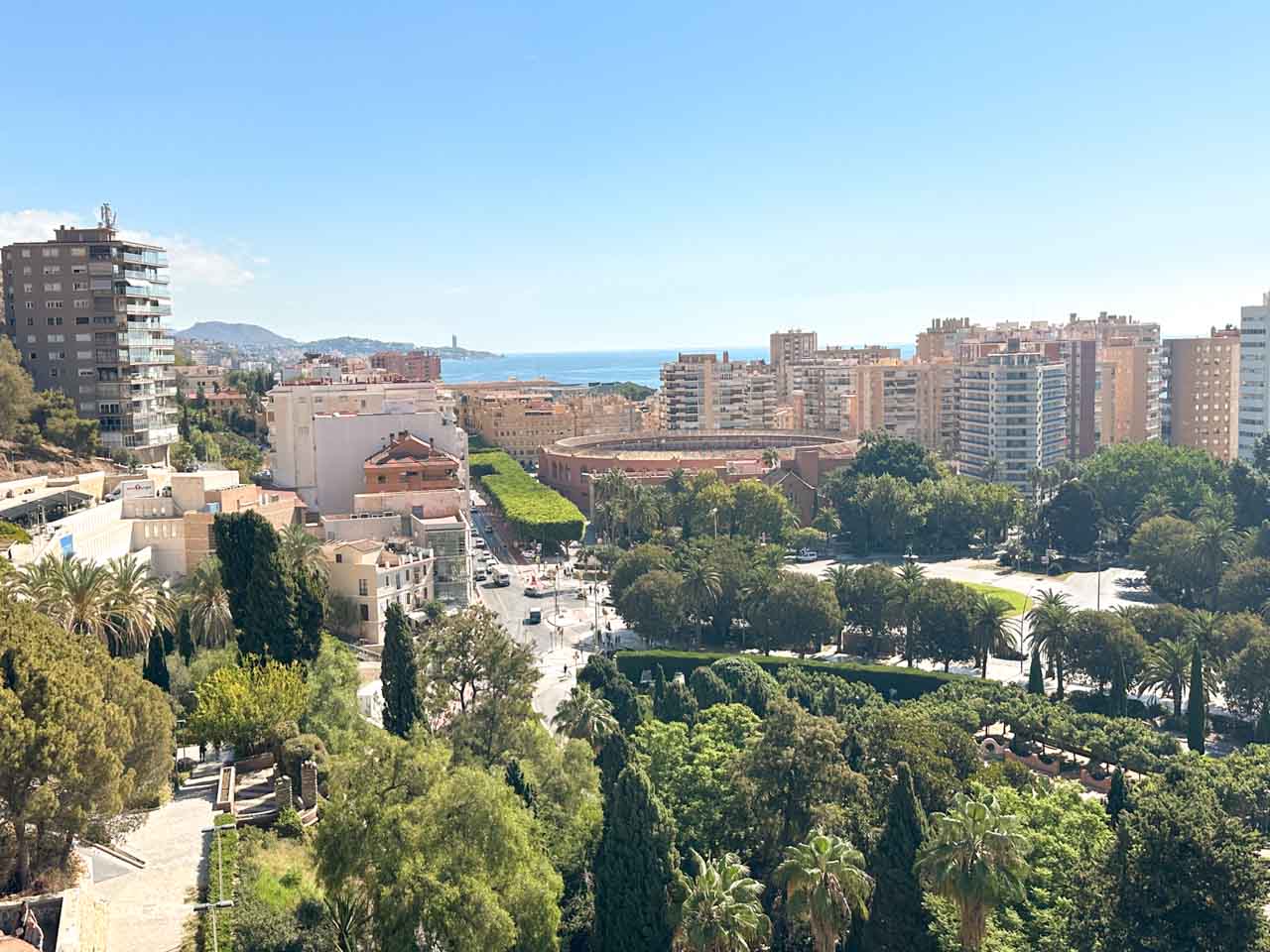 A scenic view of Malaga's cityscape seen from a hill, with green parks in the foreground and the blue sea meeting the sky in the distance