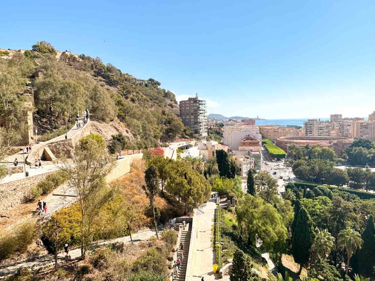 A broad path leading up to Gibralfaro Castle, with visitors walking up and down the hill, and Malaga&rsquo;s buildings and coastline in the backdrop