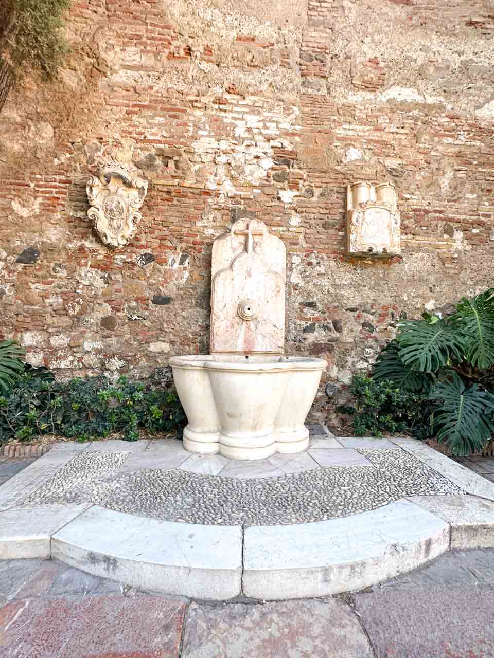 A white stone fountain against a worn brick wall with plants around it inside Alcazaba de Malaga