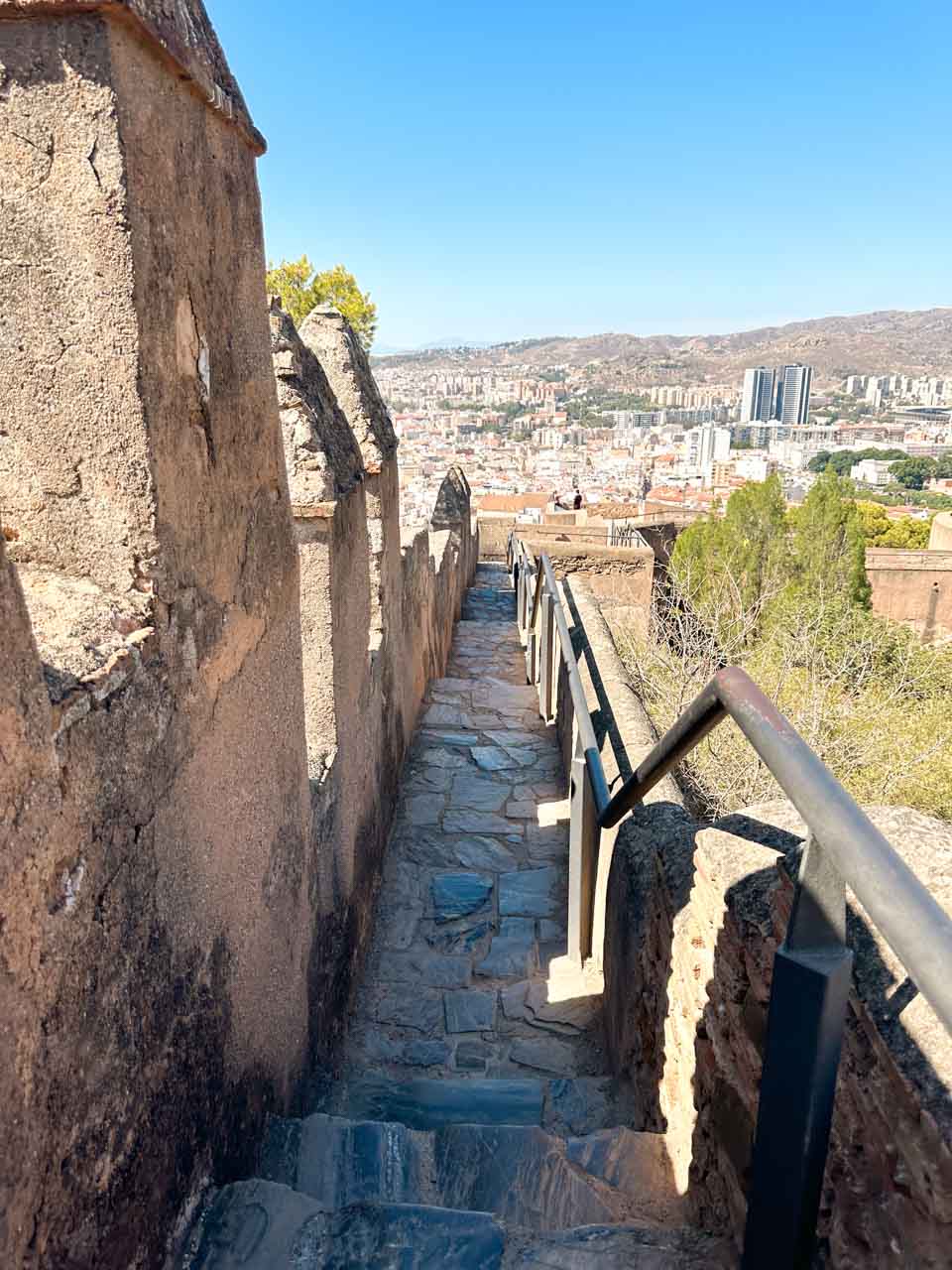 A stone stairway along the Gibralfaro fortress walls with a metal railing, overlooking the cityscape of Malaga