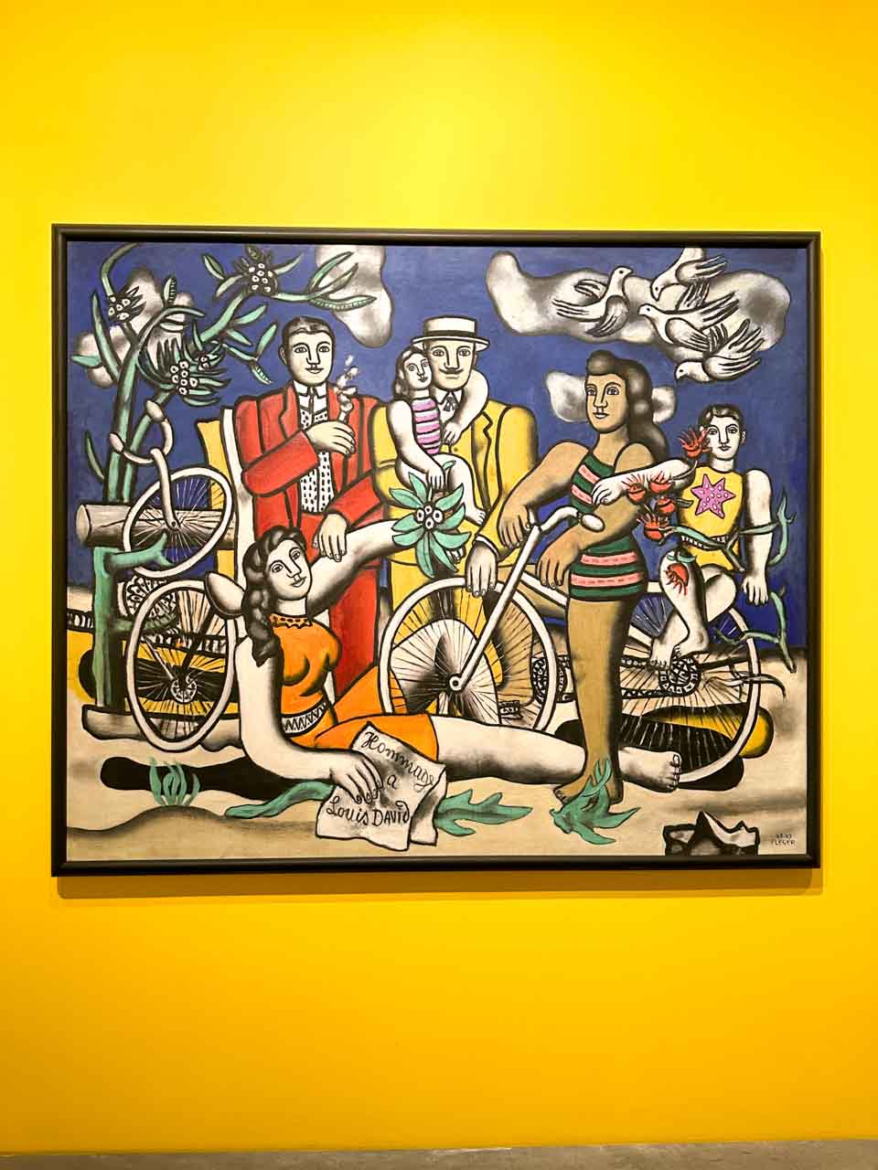 A colourful painting on a yellow wall showing a group of people with bicycles, and doves flying above them