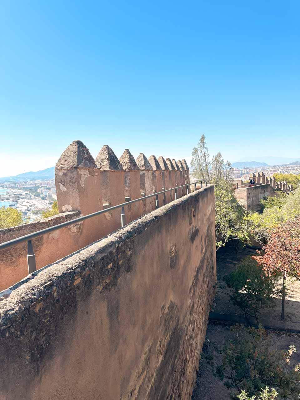 A panoramic view of a Gibralfaro Castle walkway with merlon-topped walls and the city of Malaga in the distance