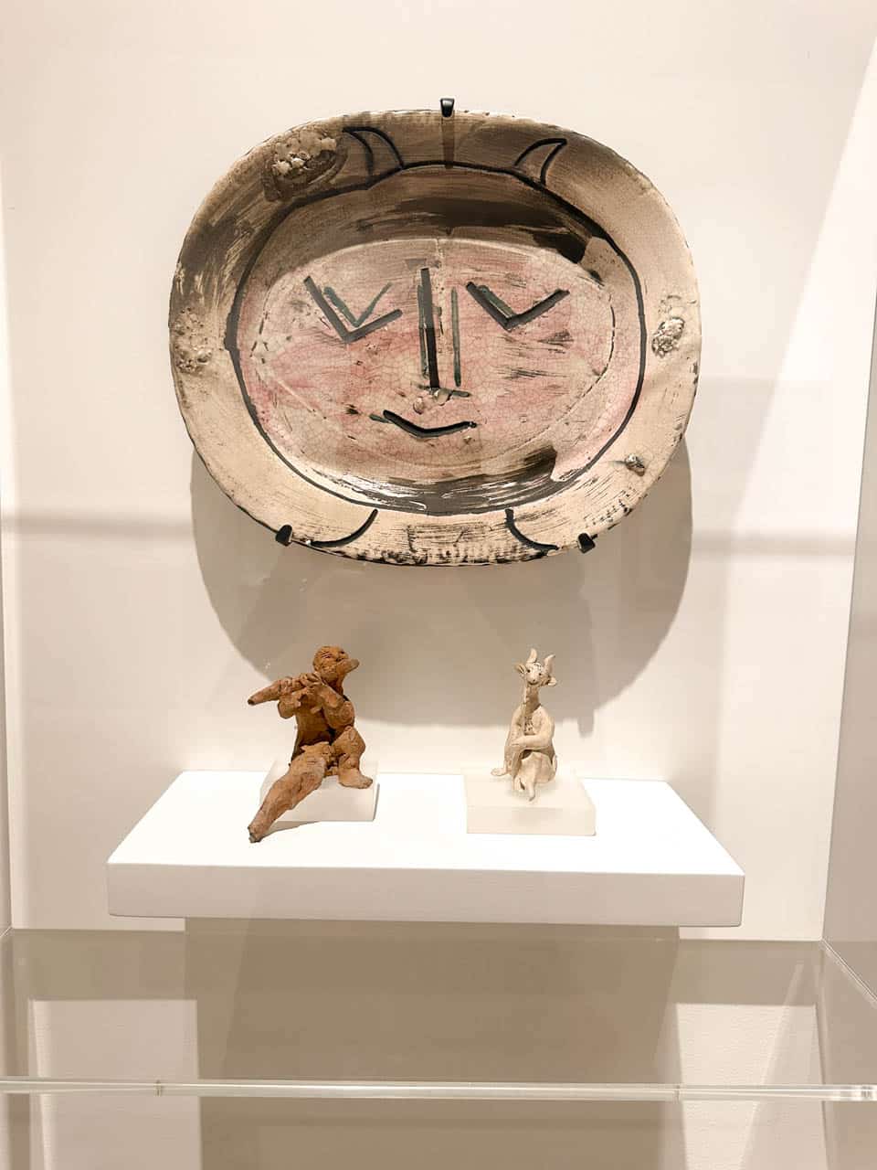 An artwork featuring a painted face on a round plate, with two small figures displayed below on a shelf