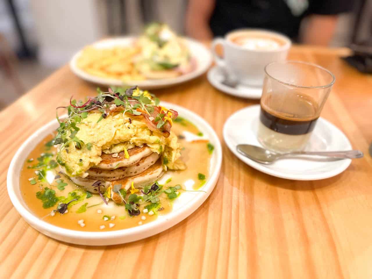 A plate with a stack of pancakes topped with scrambled eggs and garnish, beside a glass of coffee on a wooden table