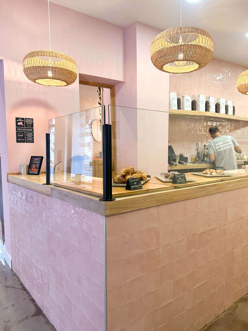 A coffee shop counter with a pink wall and wicker lampshades, where a person is making coffee