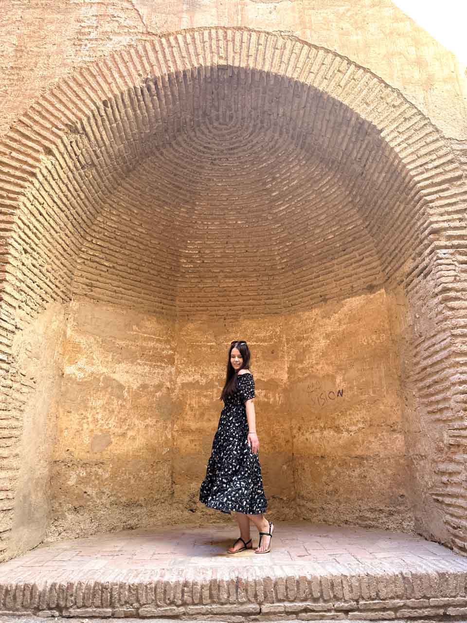 A smiling woman in a dotted dress posing in a curved, sandy-coloured nook inside the Gibralfaro Castle in Malaga