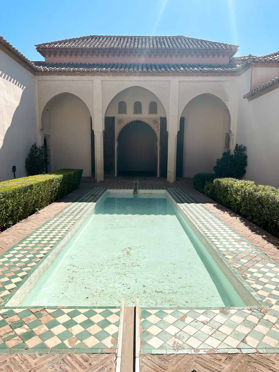 A pool in a traditional courtyard with tiled edges, surrounded by manicured hedges and archways inside the Alcazaba of Malaga
