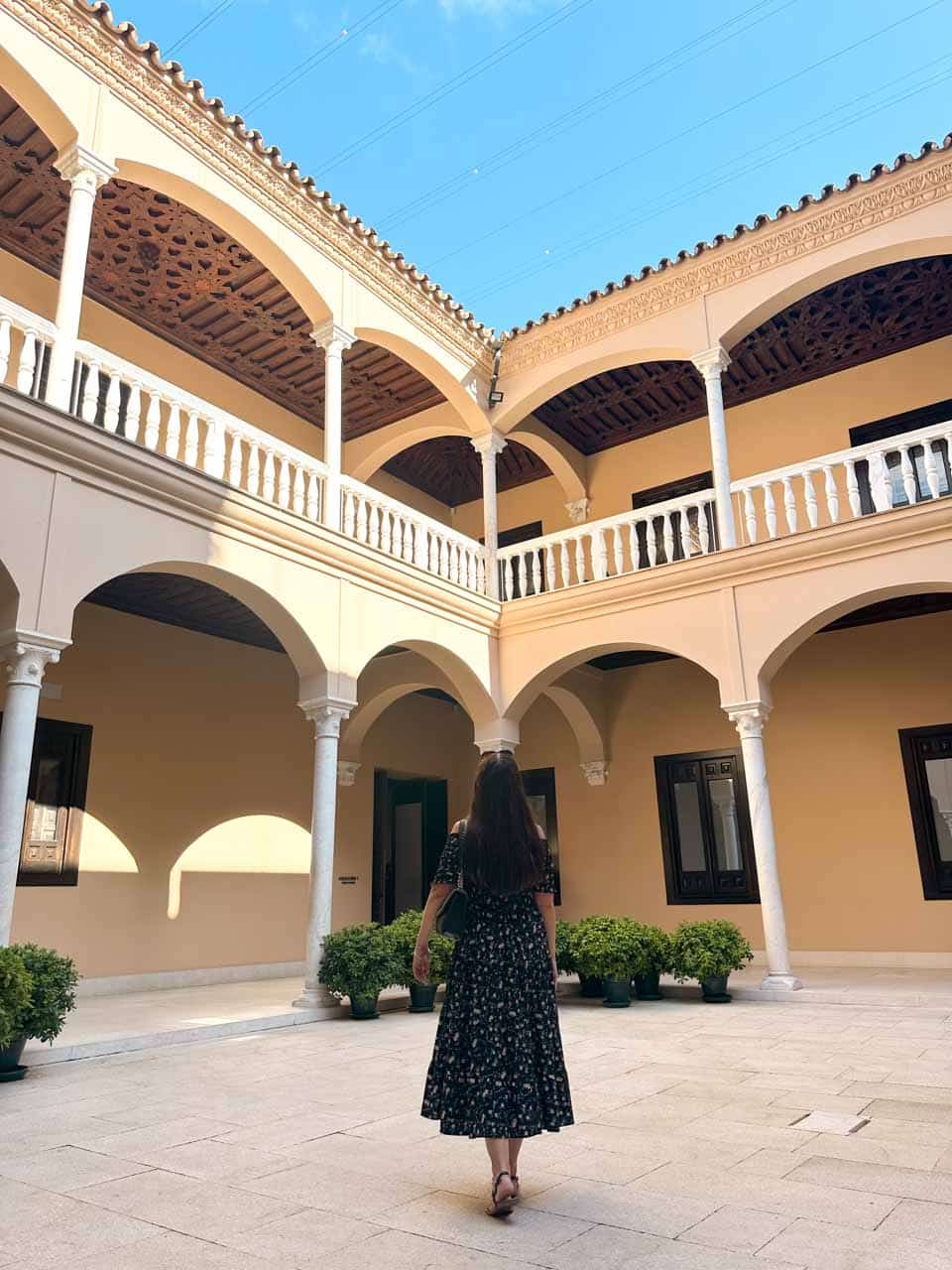 A woman standing in the courtyard of Museo Picasso in Malaga with two levels of arched balconies, cream walls, and potted plants