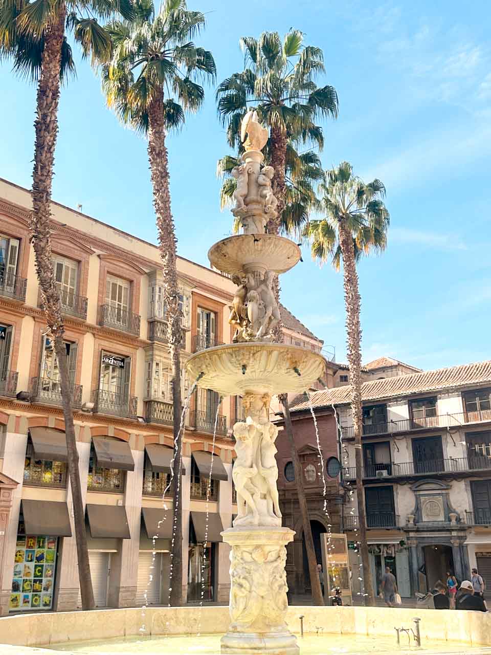 A marble fountain surrounded by palm trees, set in a square with classic buildings under a clear sky in Malaga
