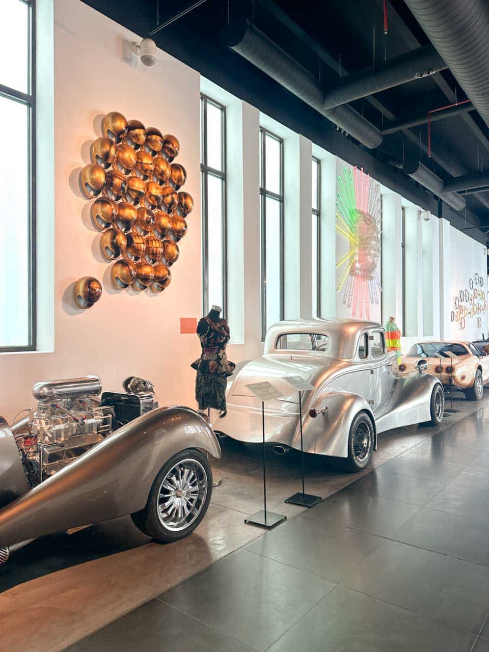 Vintage cars displayed at the Automobile and Fashion Museum in Malaga with helmet sculptures on the wall and a colourful art piece to the side