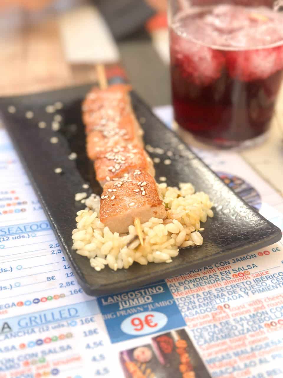 A skewered piece of tuna on a bed of rice, served on a black rectangular plate, with a blurred glass of Tinto de Verano in the background