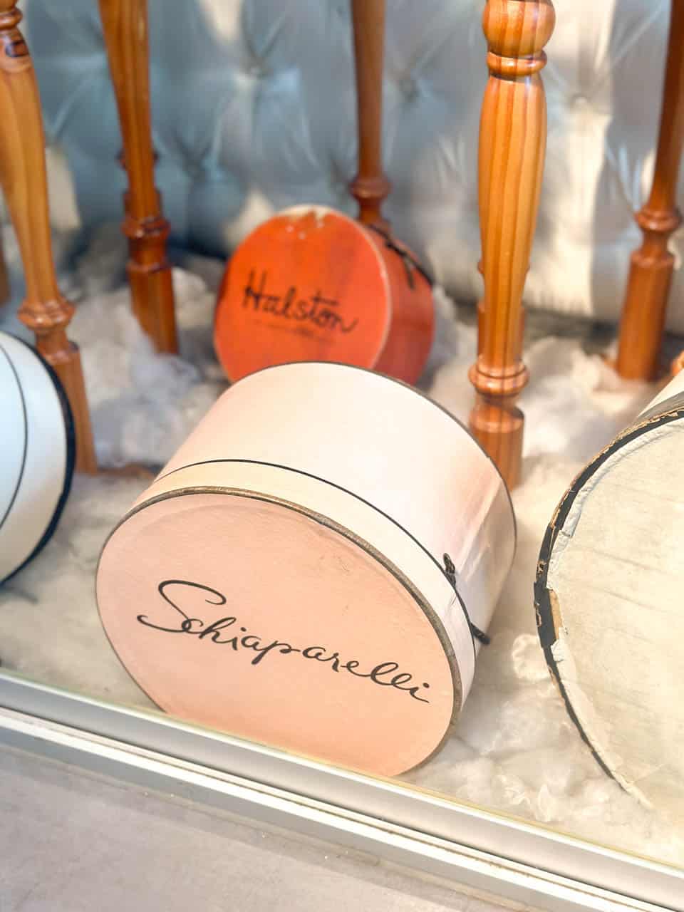 A pale pink and cream Schiaparelli hat box, set against a display of wooden chair legs and a variety of other designer hat boxes