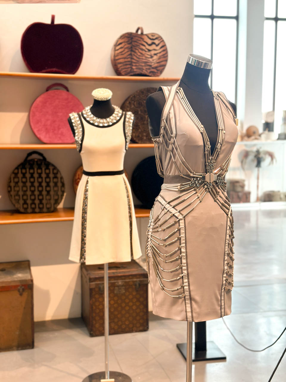 Mannequins dressed in a black and white dress and another with silver details, standing with round hat boxes on a shelf behind them