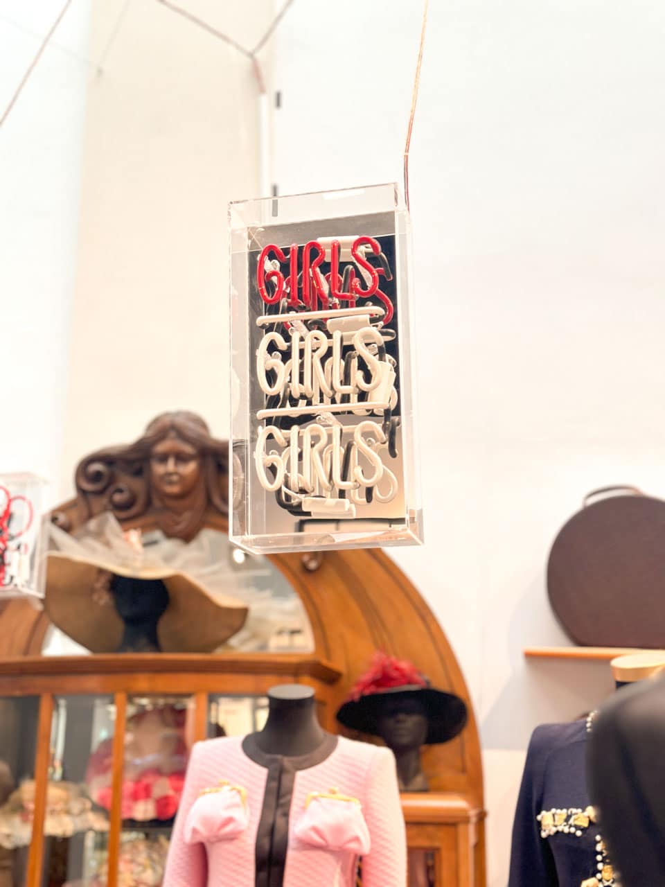 A 'GIRLS GIRLS GIRLS' neon sign in red and white, suspended above various hats and a mannequin in a pink jacket