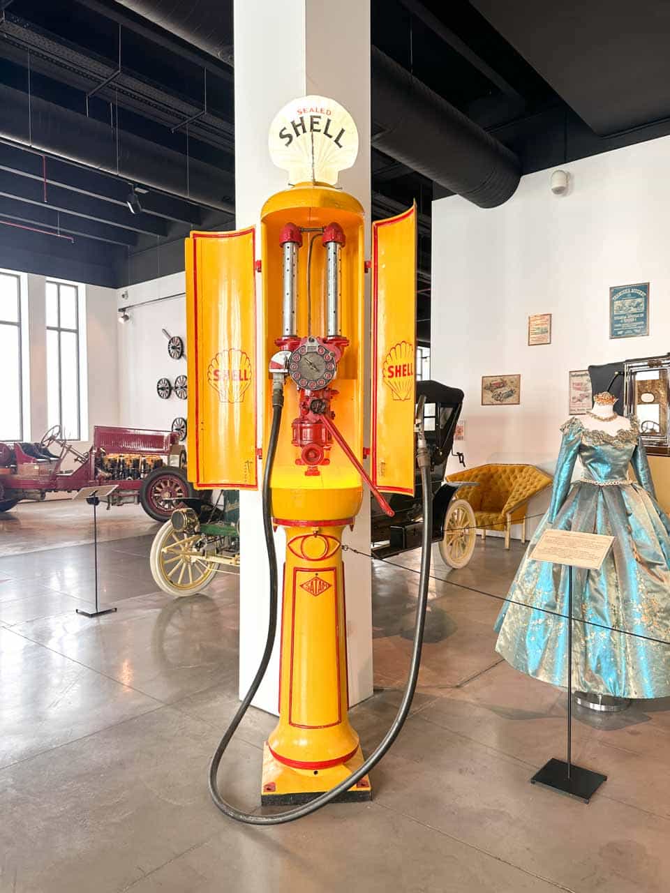 A bright yellow and red Shell petrol pump displayed in a room with vintage cars and mannequins in dresses