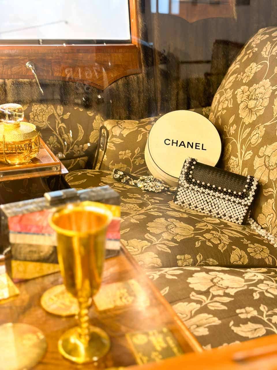 A Chanel hat box and lavish accessories inside a car with wooden panels, alongside golden glasses and shiny jewellery