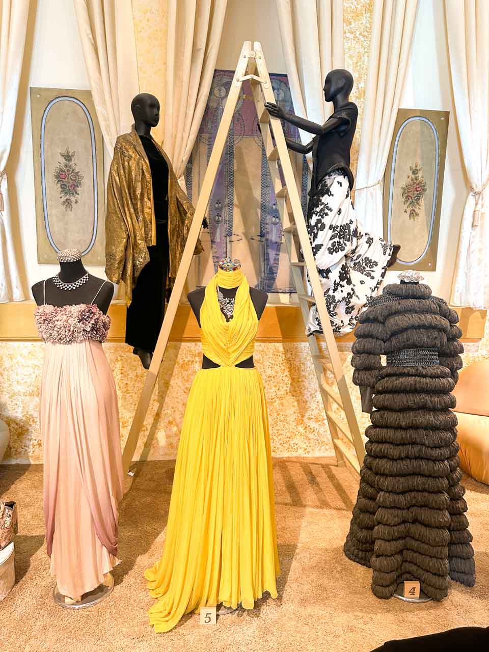 Mannequins in a fashion exhibit showcasing evening dresses, with one adjusting a painting on a ladder