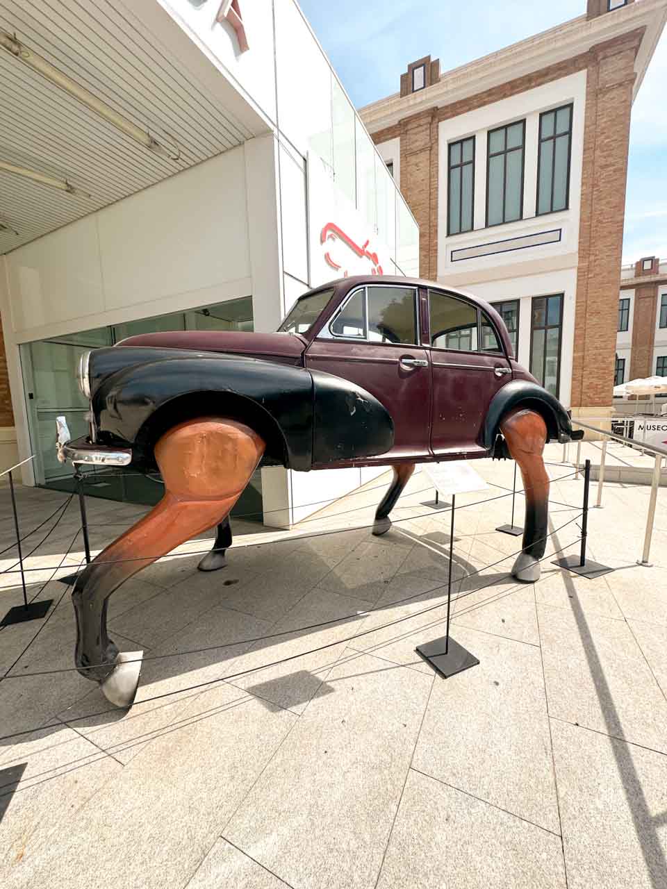 A quirky art installation featuring a classic black car with wooden horse legs, exhibited outside the Automobile and Fashion Museum in Malaga, Spain
