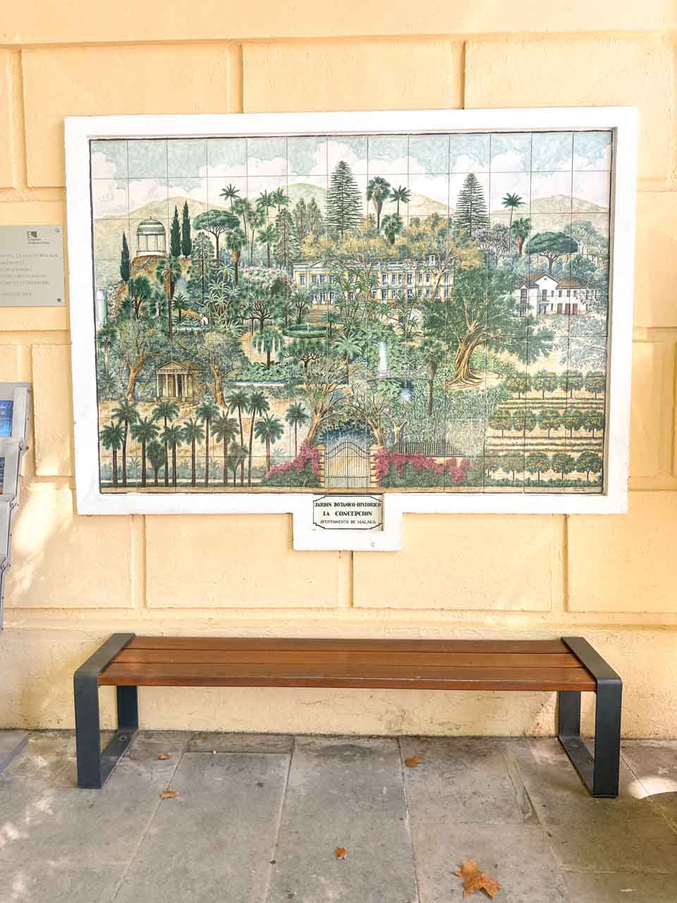 A tiled mural depicting the La Concepci&oacute;n Botanical Garden in Malaga, displayed above a simple wooden bench