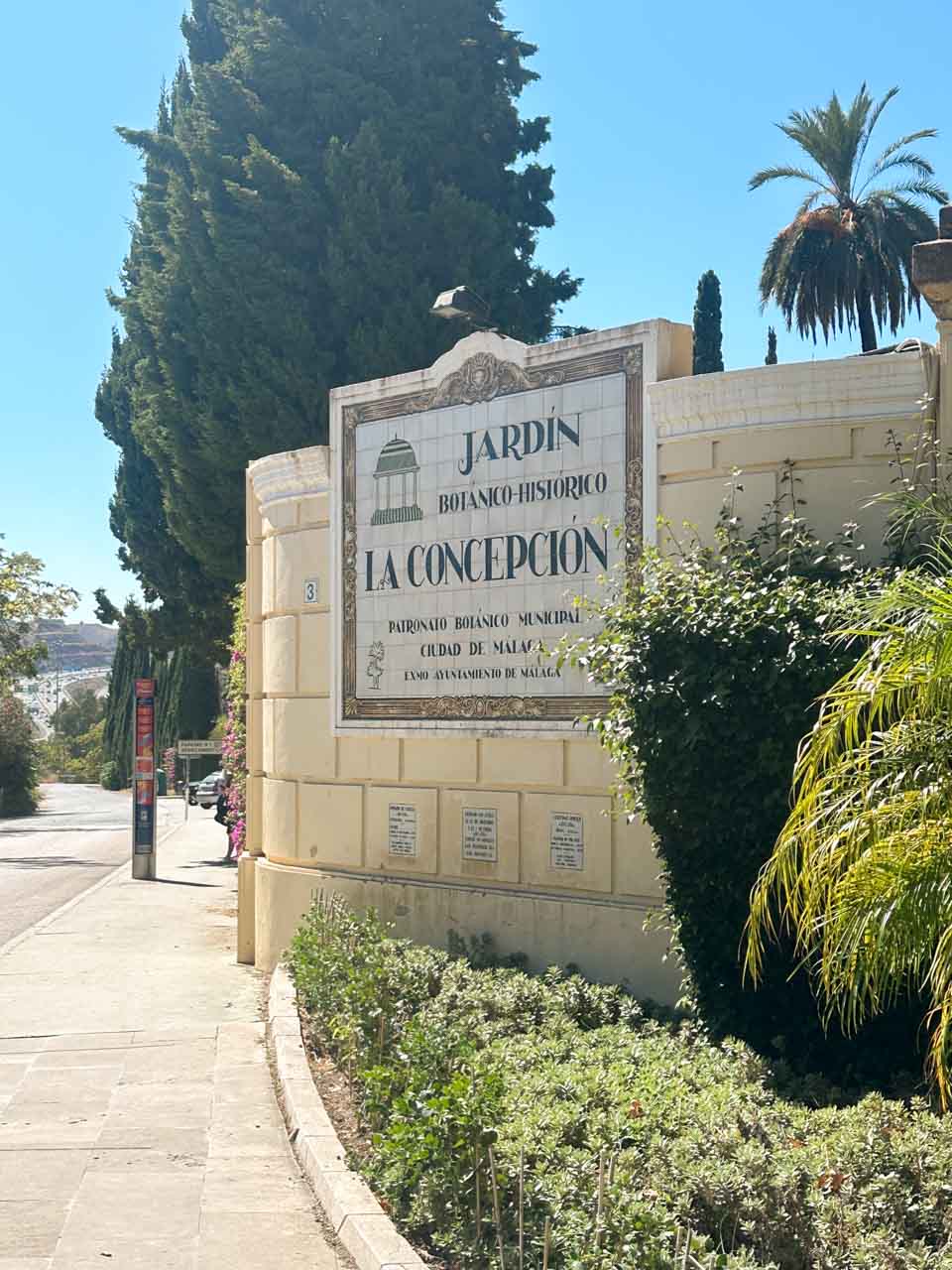 An entrance sign to the La Concepci&oacute;n Historical-Botanical Garden in Malaga, featuring decorative tiles and a backdrop of trees and palms