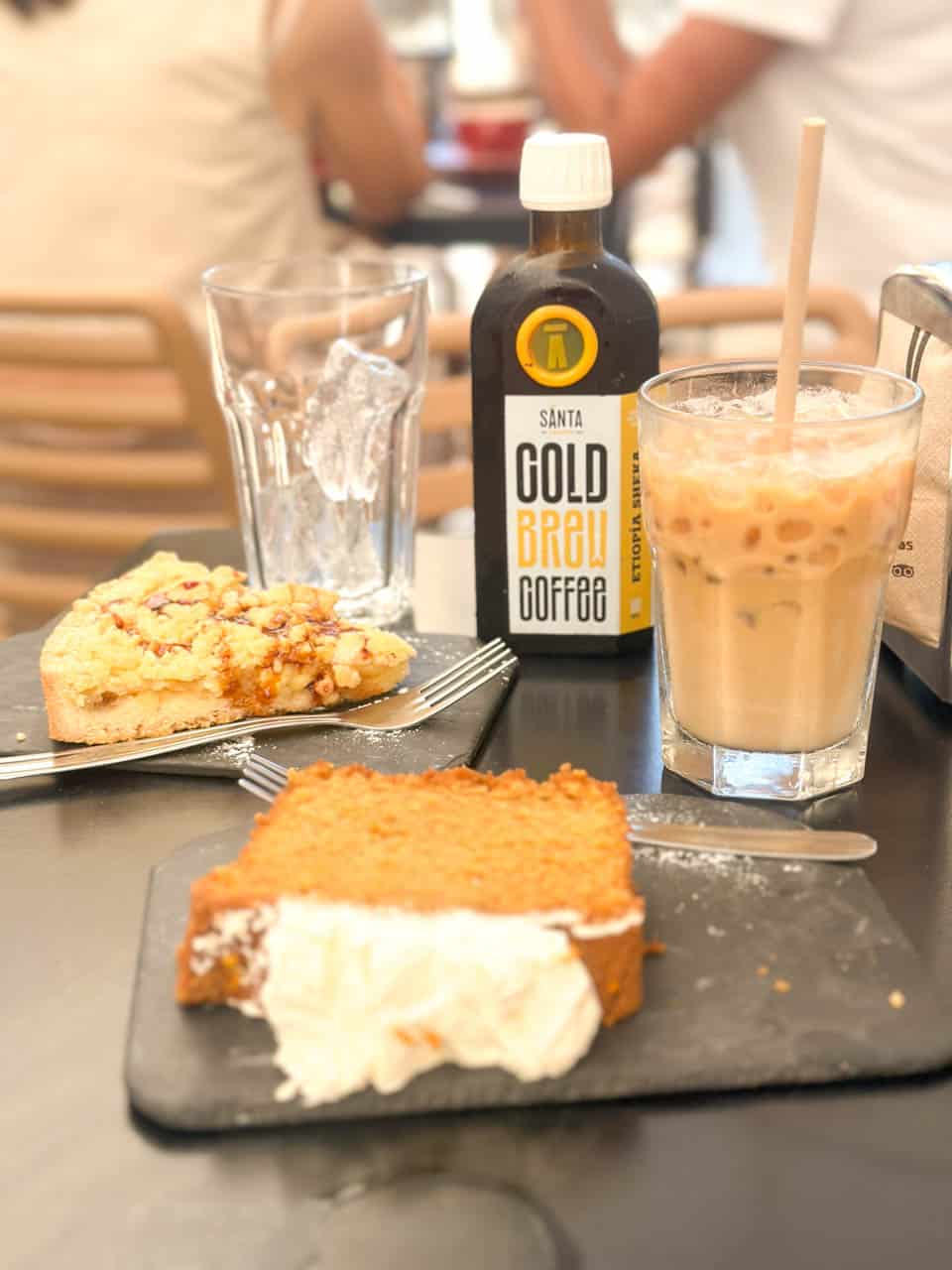A caf&eacute; table with a bottle of cold brew coffee, an iced coffee, a slice of carrot cake with cream, and a slice of crumble cake on a slate plate