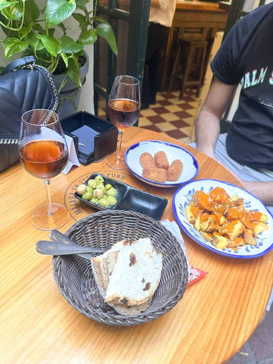 A table with glasses of sherry wine, a basket of bread, olives, and two plates of tapas, including croquettes and patatas bravas, at Bodega El Pimpi in Malaga, Spain