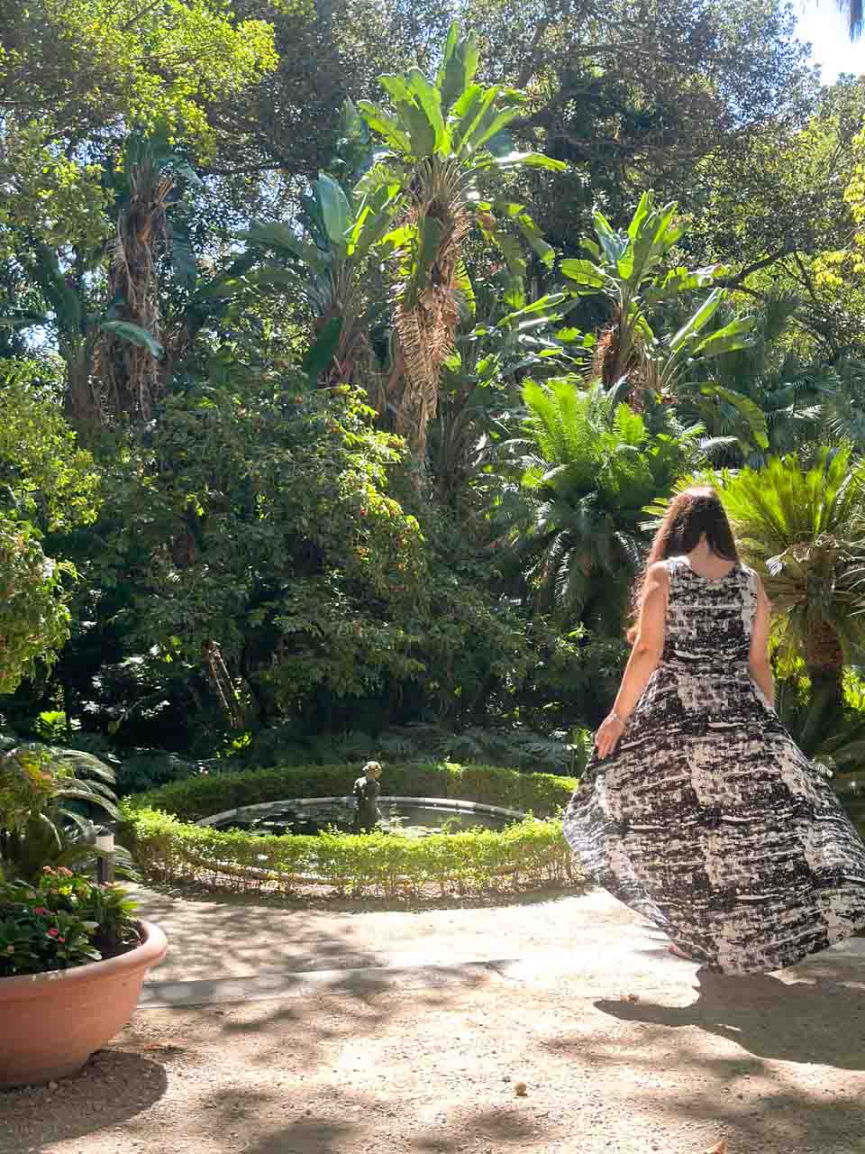 A woman in a patterned dress looking at a statue in a garden pond, surrounded by tall palms and greenery