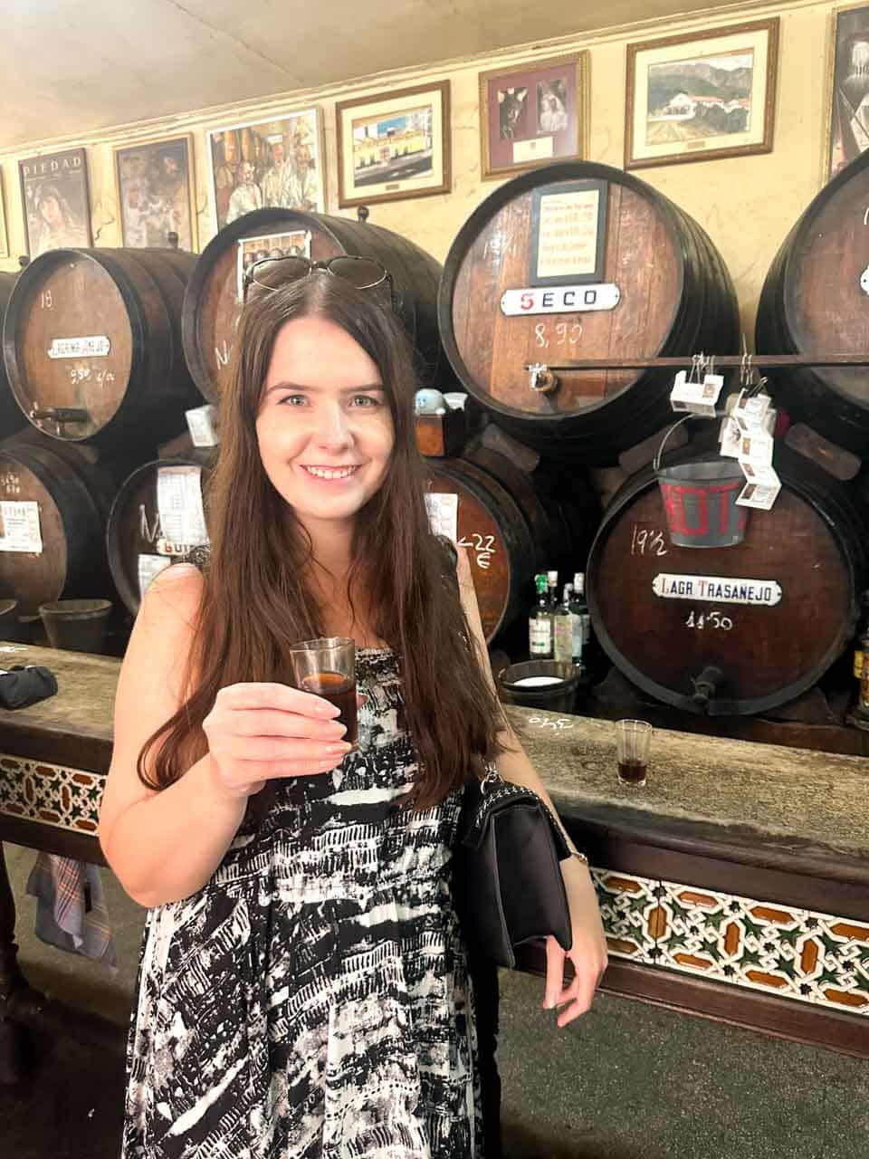A smiling woman holding a small glass of sherry in front of traditional wooden sherry barrels with prices, at Antigua Casa de Guardia in Malaga, Spain