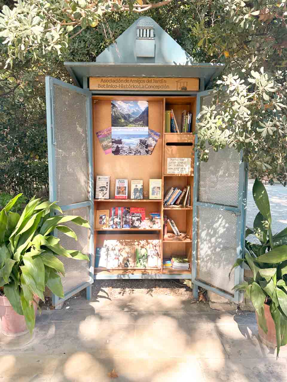 A blue book exchange booth filled with books, under the shade of a tree in a park in Malaga, Spain