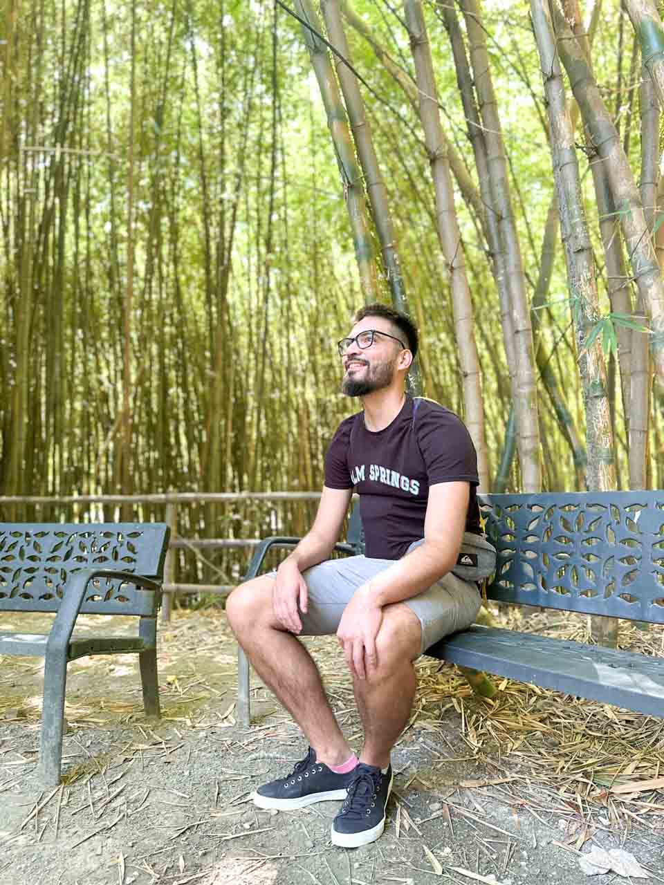 A man smiling and sitting on a metal bench in a bamboo garden, with tall bamboo shoots in the background