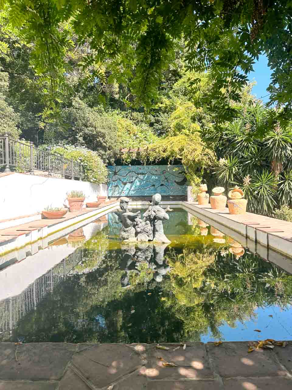 A statue in the middle of a reflective pond, surrounded by trees and potted plants inside Malaga's botanical gardens