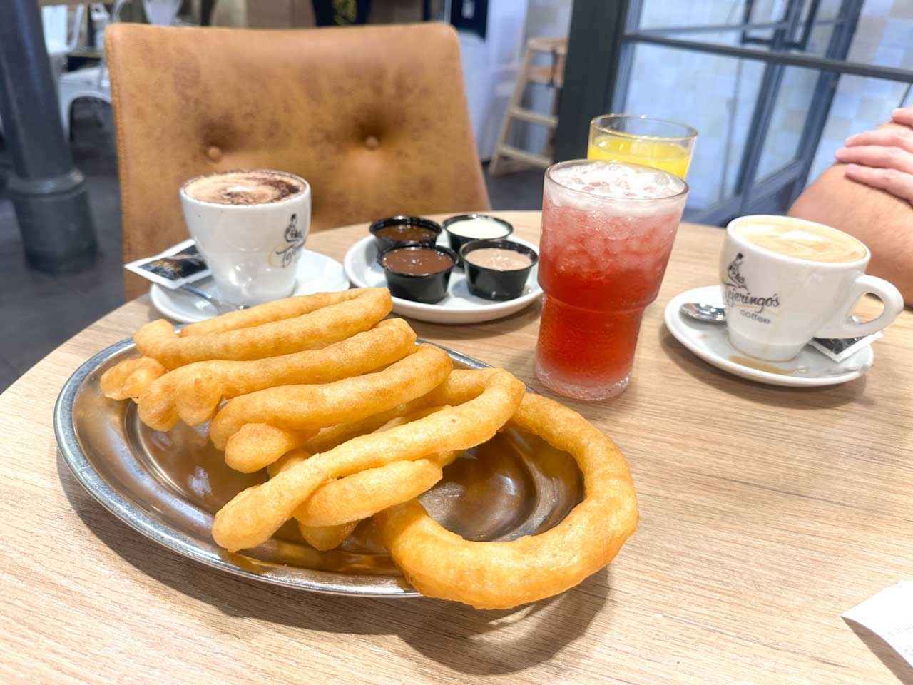 A plate of churros with four dipping sauces, two cups of coffee and glasses of drinks on a café table at Tejeringo’s Coffee