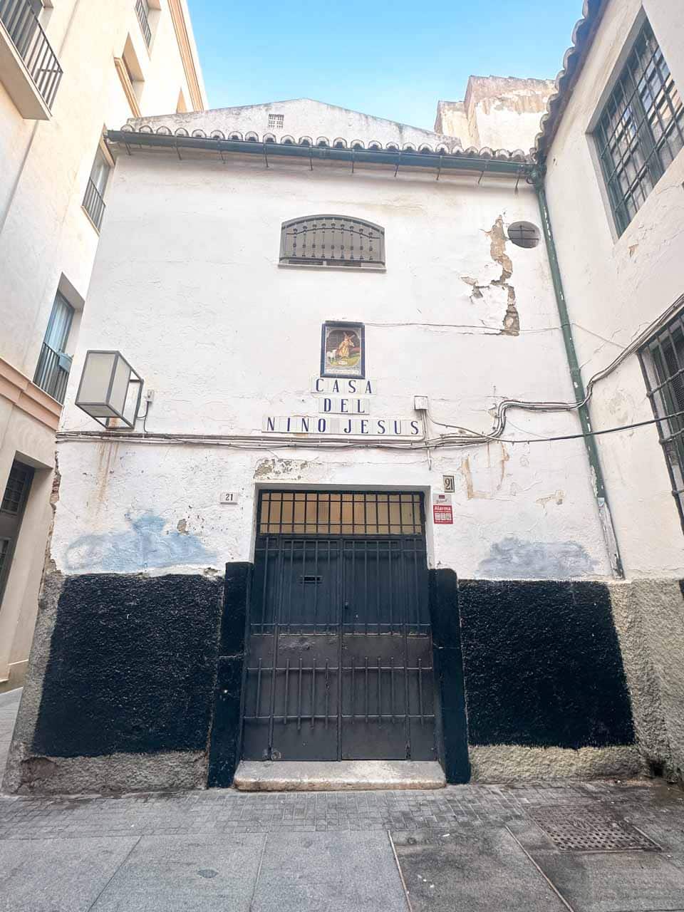 The exterior of Casa del Ni&ntilde;o Jes&uacute;s, a white building with religious signage and black and grey lower walls, in a narrow street in Malaga