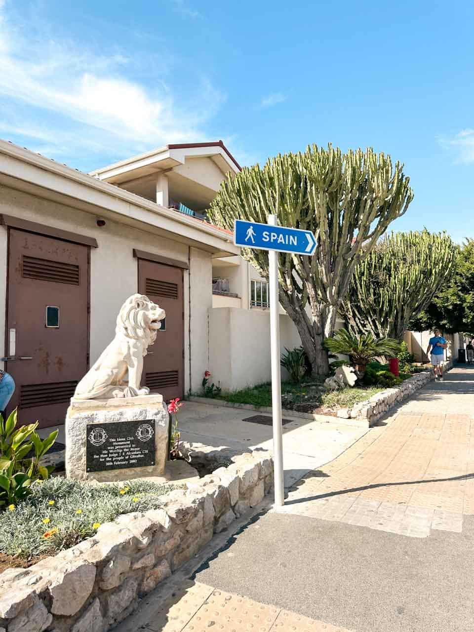 A directional sign pointing towards Spain next to a statue of a white lion, surrounded by greenery