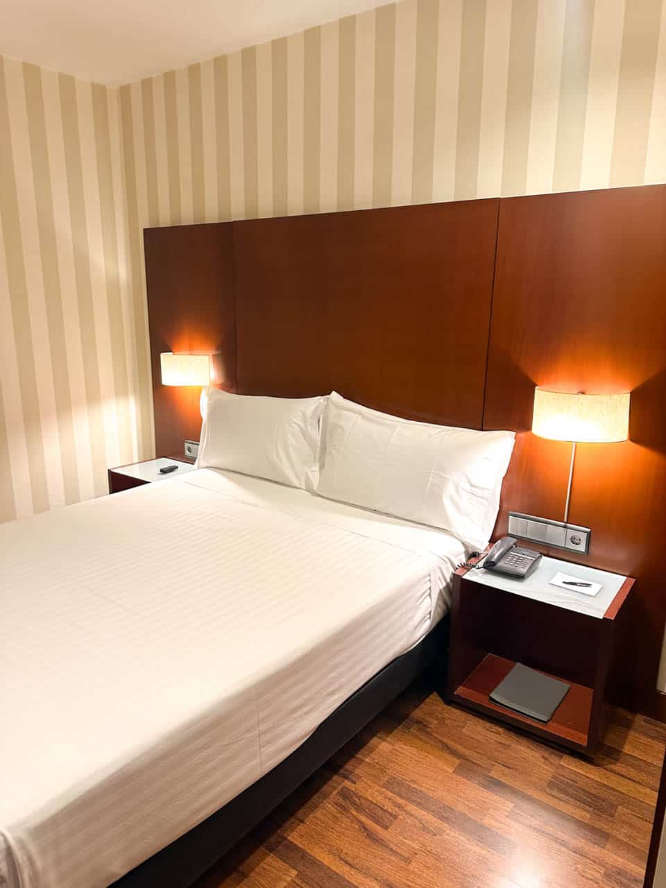 A cosy hotel room featuring a large double bed with white linens, flanked by two bedside tables with lamps, against a striped cream and beige wall