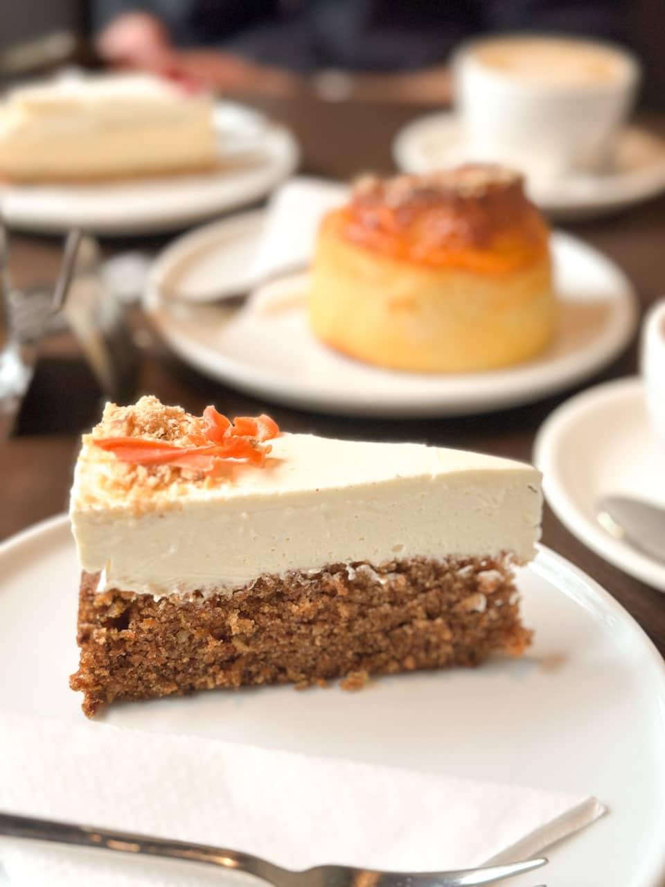 A close-up of a slice of carrot cake on a table, with a slice of cheesecake and a savoury bun in the background